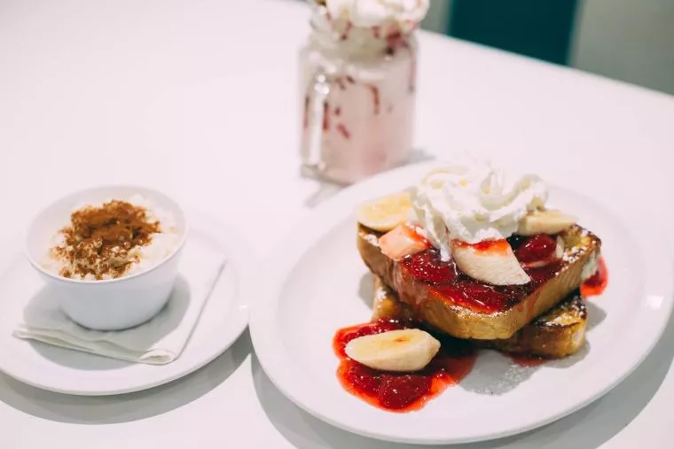A plate with French toast topped with sliced bananas, strawberries, strawberry sauce, and whipped cream sits beside a cup of rice pudding and a glass of a pink milkshake topped with whipped cream.