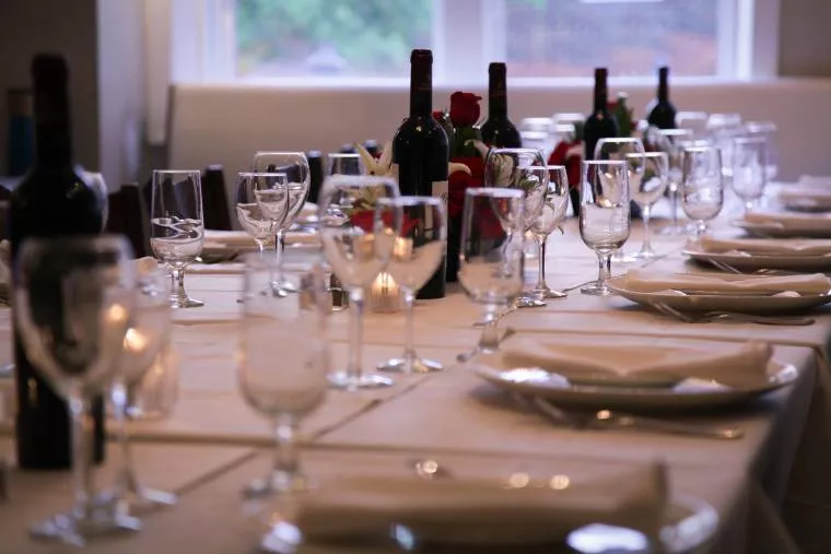 A formal dining table set with white tablecloth, plates, wine glasses, and several bottles of red wine, arranged for a special event or dinner party. Roses and candles create an elegant atmosphere.