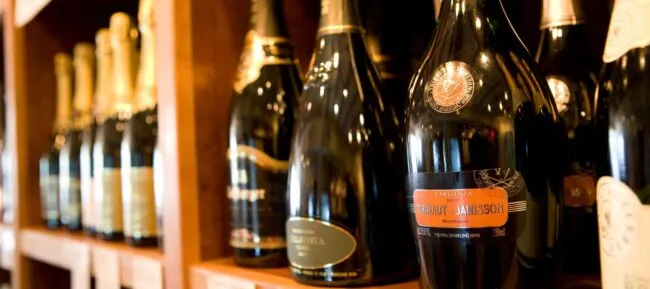Bottles of sparkling wine and champagne are displayed on wooden shelves in a shop, with labels and foil tops visible, creating an elegant and inviting atmosphere.
