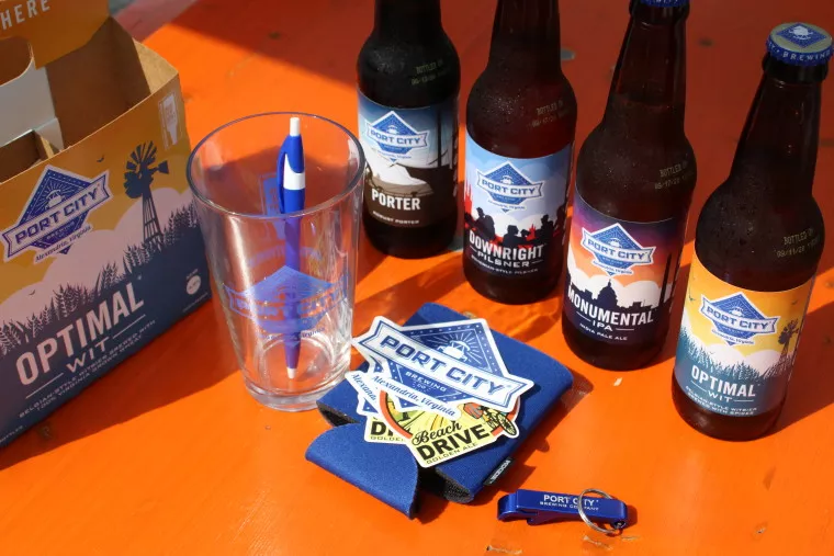 Four Port City beer bottles, an empty branded glass with a blue pen, a cardboard beer carrier, Port City stickers, a blue bottle opener, and blue coasters are arranged on an orange table.