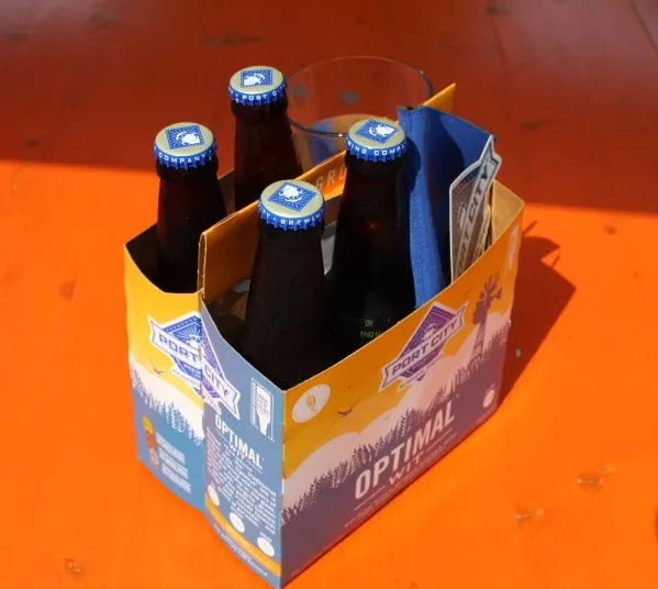 A four-pack of bottled beer with blue caps in a cardboard carrier labeled Optimal, placed on an orange wooden surface. A glass is partially visible behind the bottles.