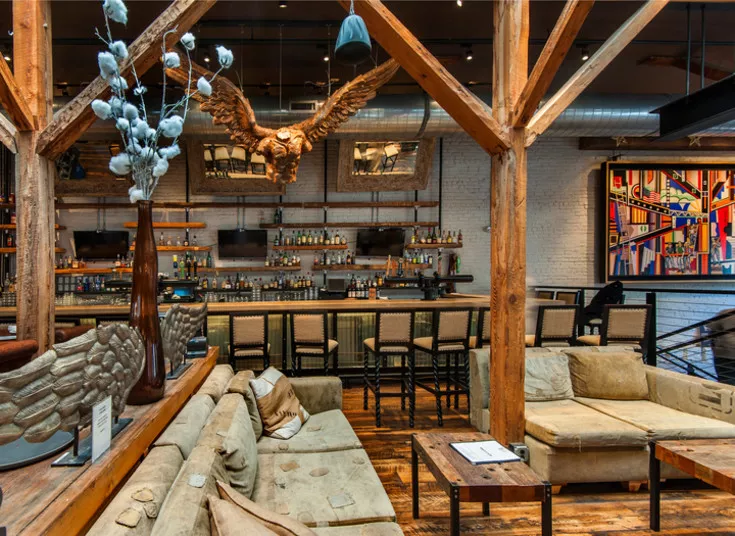 Cozy lounge area with tan sofas and wood accents in front of a bar. Wooden beams cross the ceiling, an eagle sculpture hangs above, and a colorful abstract painting decorates a white brick wall.