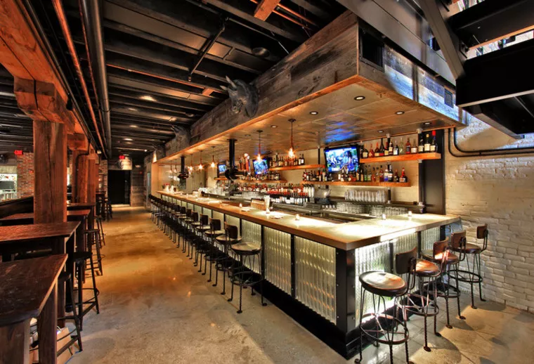 A modern bar with high stools, a polished counter, exposed beams, shelves of bottles, flat-screen TVs, and warm lighting creating an inviting atmosphere.
