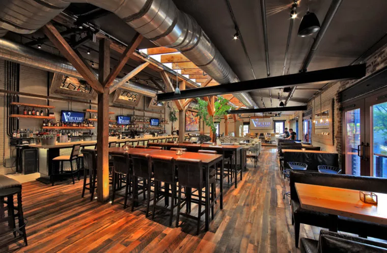 Industrial-style restaurant interior with wooden floors, exposed beams, metal ductwork, high tables with stools, a long bar area, multiple TV screens, and large windows letting in natural light.