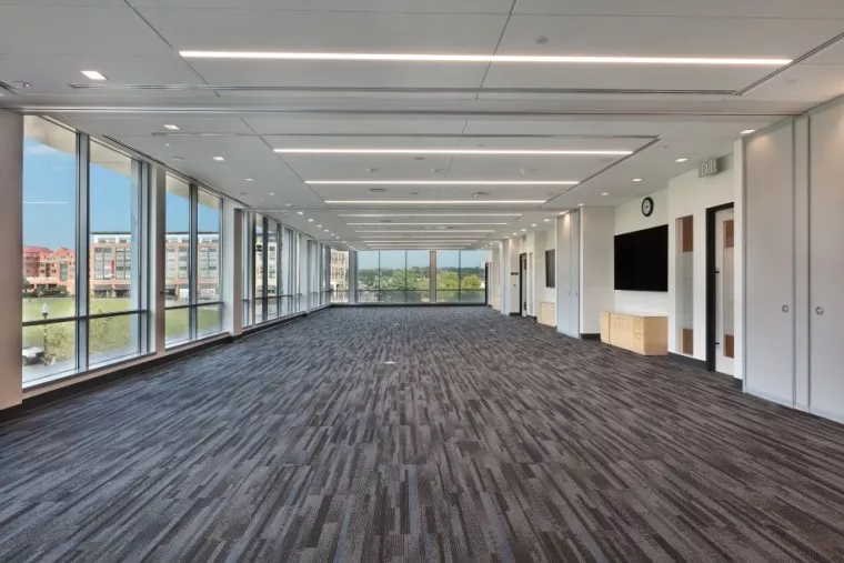 A large, modern, empty conference room with carpeted floors, floor-to-ceiling windows on two sides, white walls, and a wall-mounted TV. Natural light fills the space and buildings are visible outside.