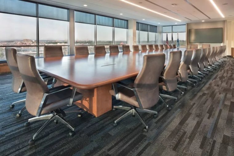 A spacious, modern conference room with a large wooden table surrounded by many empty swivel chairs. Large windows provide natural light and a cityscape view. A flat-screen monitor is on the wall.