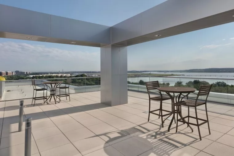 A spacious rooftop terrace with tall metal frames, two round tables with chairs, glass railing, and a scenic view of a river, greenery, and city buildings in the distance under a partly cloudy sky.