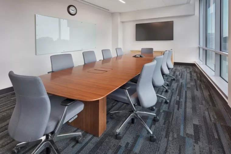 Modern conference room with a long wooden table, ten gray office chairs, a wall-mounted TV, a whiteboard, a clock, and large windows letting in natural light. The room is empty and well-lit.