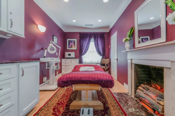A cozy spa room with purple walls, a massage table covered in a red sheet, a decorative rug, fireplace, medical equipment, cabinets, and a window with purple curtains letting in natural light.