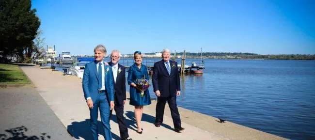 Four people dressed formally walk along a riverside path on a sunny day. Two men wear blue suits, one man wears a dark suit, and a woman in a teal dress holds a bouquet. Boats and trees are visible in the background.