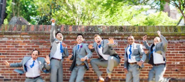 Six men in light gray suits and blue ties jump enthusiastically in front of a brick wall and greenery, smiling and making energetic gestures, as if celebrating or posing for a fun group photo.