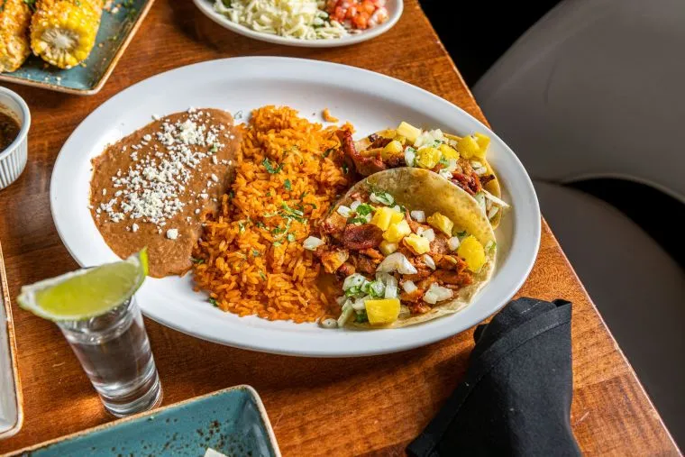 A plate with two tacos filled with meat, pineapple, onions, and cilantro, served with Mexican rice and refried beans. A glass of water with a lime wedge is nearby on a wooden table.