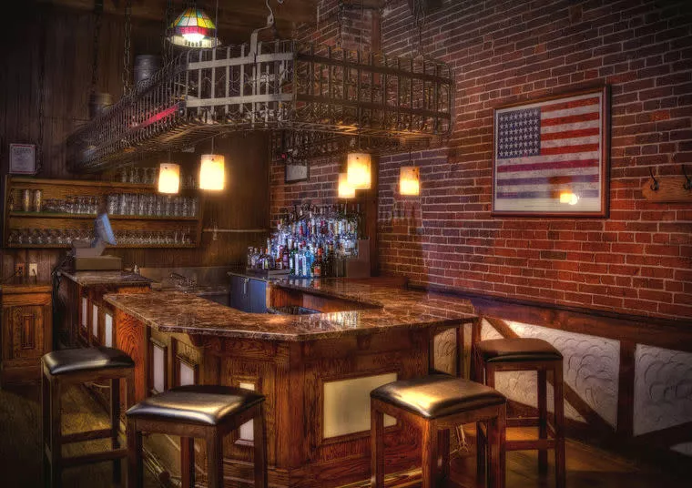 A cozy bar with a marble countertop, wooden stools, shelves of glasses, a hanging rack above, and an American flag on a brick wall, softly lit by warm pendant lights.