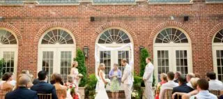 A bride and groom stand facing each other during an outdoor wedding ceremony in front of a brick building, with guests seated and wedding party members standing on either side.