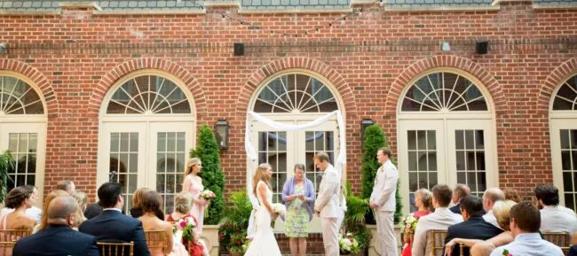 A bride and groom stand facing each other during an outdoor wedding ceremony in front of a brick building, with guests seated and wedding party members standing on either side.