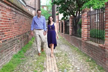 A man and woman hold hands while walking down a cobblestone alley lined with brick walls and iron fences. Both are smiling and casually dressed, with greenery along the path and trees in the background.
