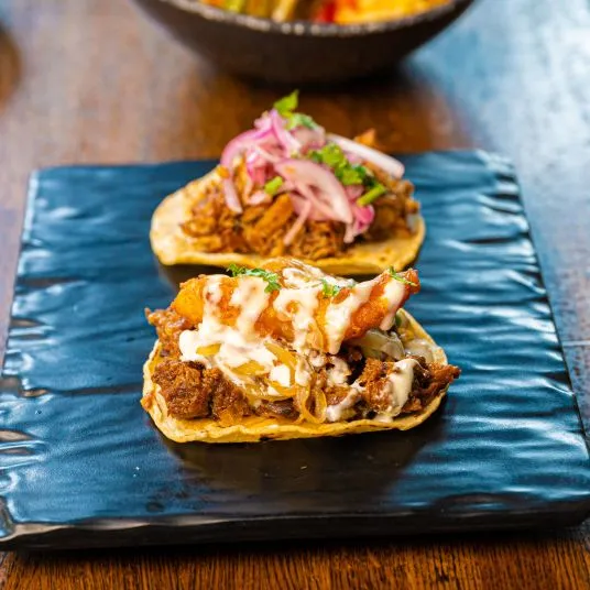 Two tostadas on a black rectangular plate; one topped with pulled meat, pink pickled onions, and herbs, the other with meat, fried shrimp, sautéed onions, and drizzled creamy sauce.