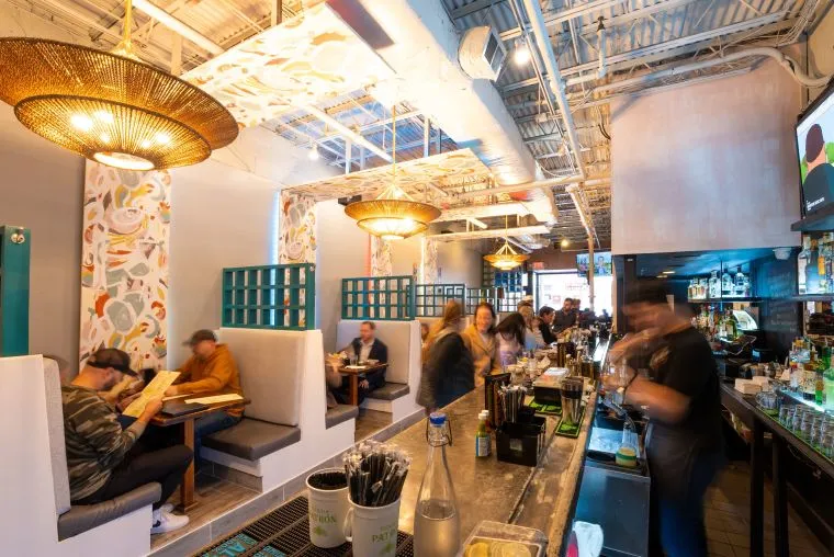 A vibrant restaurant interior with modern decor, booths along the wall, and a long bar in the foreground. Patrons are dining and staff are moving behind the bar, creating a lively atmosphere. Warm lighting hangs from the ceiling.
