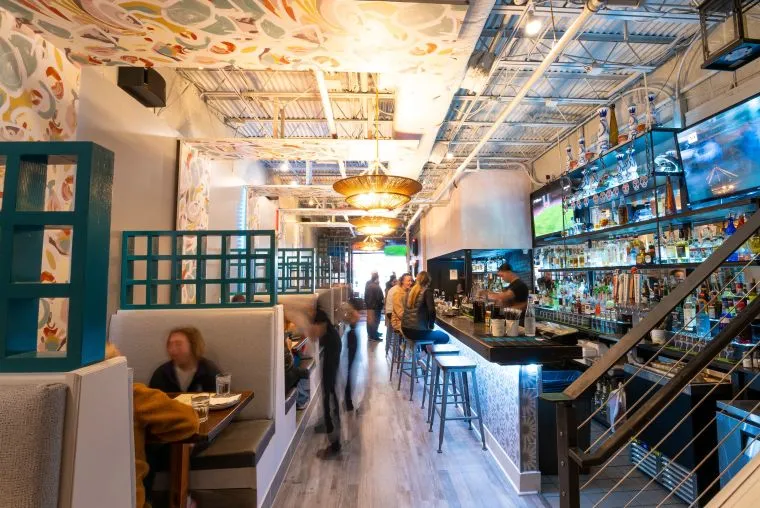 A modern bar and restaurant with colorful decor, booth seating on the left, a long bar on the right, and large mirrors reflecting bottles and a TV. People are dining, drinking, and moving through the bright space.