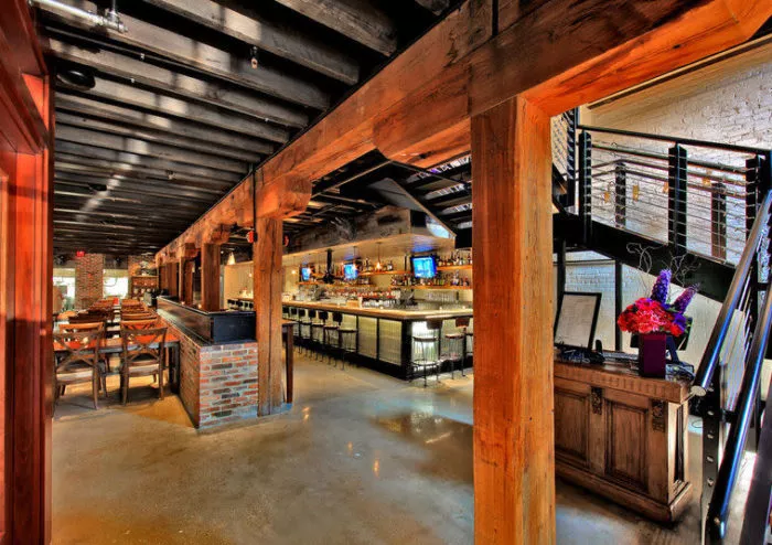 Modern bar and restaurant interior with exposed wooden beams, brick accents, a long bar with stools, tables and chairs, and a staircase beside a wooden host stand with a bouquet of flowers. Warm lighting creates a cozy atmosphere.