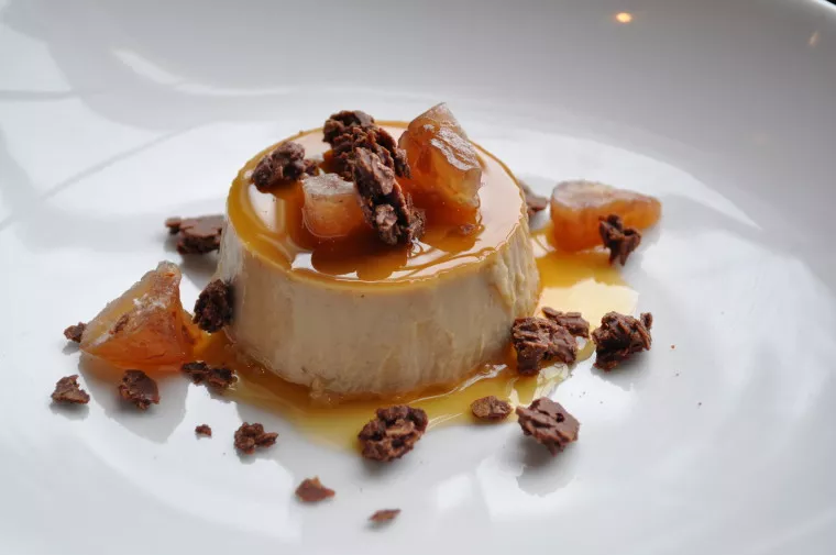 A creamy flan topped with caramel sauce, chocolate flakes, and chunks of candied fruit, served on a white plate.