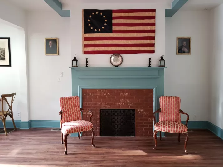 A Colonial-style room with a brick fireplace, two patterned armchairs, and a large Betsy Ross American flag hanging above the mantel, flanked by framed portraits and two lanterns. Sunlight streams in from the left.