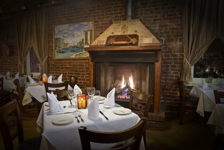 A cozy restaurant dining area with white tablecloths, elegant place settings, and a lit fireplace set in a brick wall, with curtains and a cityscape painting on the wall.