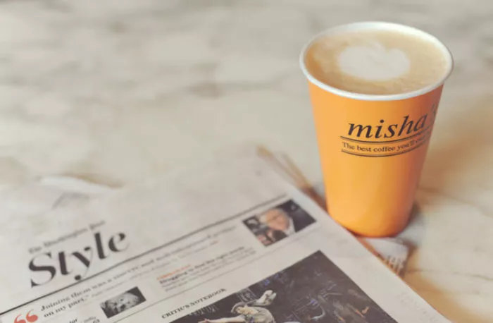 A yellow takeaway coffee cup with latte art sits on a marble surface next to a folded newspaper section titled Style.