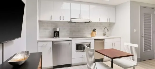 Modern kitchen with white cabinets, stainless steel appliances, a coffee maker, a knife block, a sink, and a small wooden dining table with two white chairs. A TV and decorative bowl are also visible.