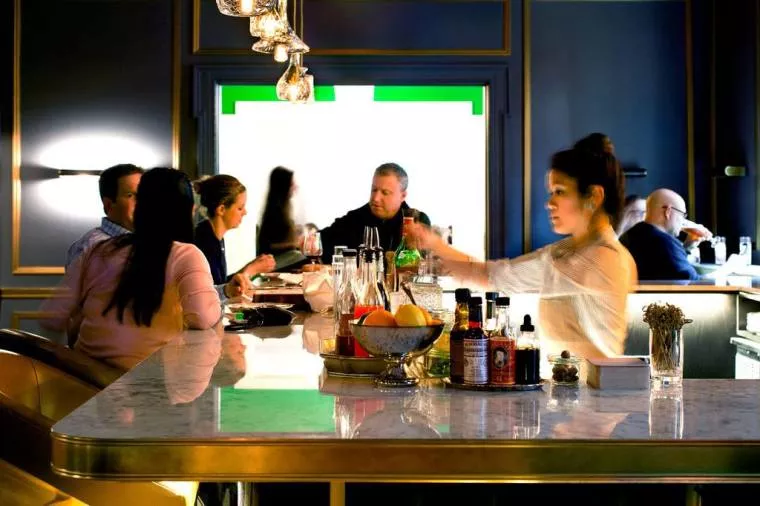 People sit and talk around a marble bar counter in a modern restaurant with dark walls. Bottles, fruit, and condiments are on the counter. A woman in white serves drinks while warm lights hang above.