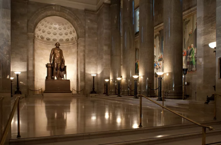 A grand marble hall features a large bronze statue of George Washington at the far end, surrounded by tall columns, wall murals, and lit by elegant lamps, creating a formal and historic atmosphere.
