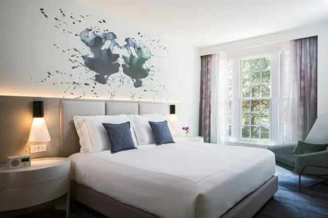 A modern hotel room with a large bed, two bedside tables with lamps, abstract art above the headboard, a window with sheer curtains, and a green armchair in the corner.