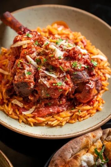 A braised lamb shank topped with tomato sauce and herbs, served over a bed of orzo pasta on a beige plate. Flakes of cheese are sprinkled on top. A slice of bread is partially visible on the side.