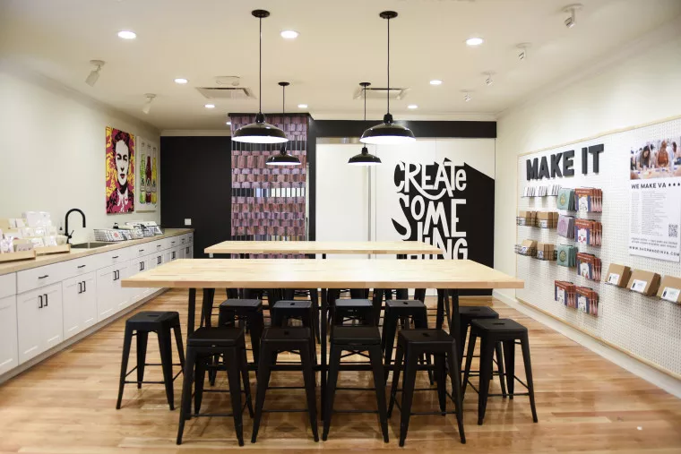 A modern, well-lit art studio features wooden tables with black stools, white cabinets, wall art, and a display of cards. A large mural reads “CREATE SOMETHING.” Pendant lights hang from the ceiling.