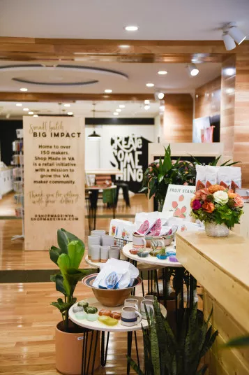 A modern shop interior with wooden floors and shelves displays colorful mugs, plates, towels, and flowers. A wooden sign highlights local makers, and a “CREATE SOMETHING” mural is visible in the background.