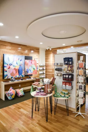 A modern gift shop with wooden floors, colorful abstract artwork on the wall, display tables with candles and books, and shelves stocked with various items and greeting cards.