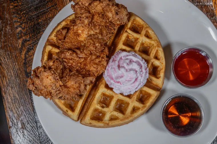 A plate of golden waffles topped with a scoop of pink whipped cream and crispy fried chicken, served with two small cups of syrup on the side.