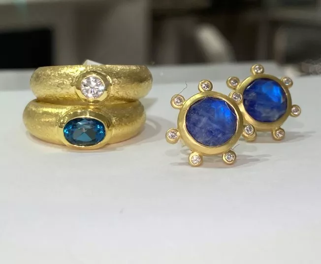 Two gold rings, one with a clear round gemstone and the other with an oval blue gemstone, displayed next to a pair of gold earrings with large blue stones surrounded by small clear gems.