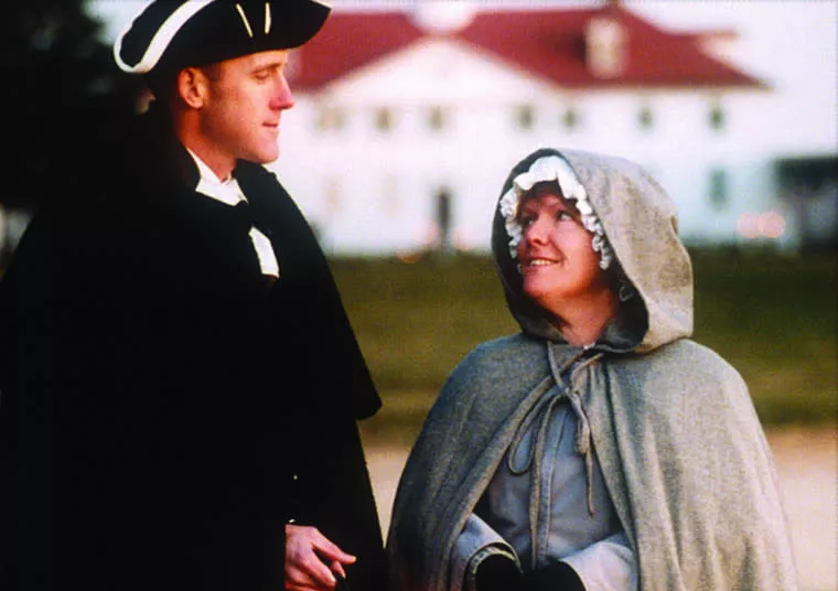 A man in colonial attire and a woman in a gray bonneted cloak stand outdoors, looking at each other and smiling, with a large white building and greenery blurred in the background.