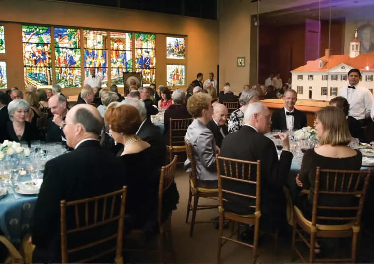 Guests dressed formally are seated at round tables set for dinner in an elegant room with stained glass windows and a large model house display. The atmosphere appears festive, with people engaged in conversation.
