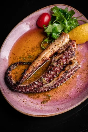 A grilled octopus tentacle served on a pink plate with arugula, a lemon wedge, a cherry tomato, and a pool of seasoned olive oil.