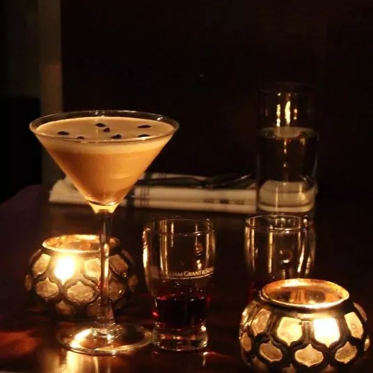 A martini glass filled with a creamy cocktail, garnished with coffee beans, sits on a dark table surrounded by candles, a shot glass with a dark drink, and a glass of water in the background.