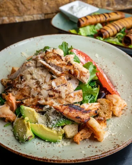 A plate of grilled chicken salad with sliced avocado, croutons, greens, cheese, and shrimp, garnished with shredded onions. In the background, a plate of grilled wraps with a small bowl of dipping sauce.