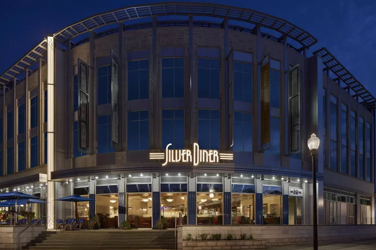 A modern, round-front building at dusk with large windows and a bright neon sign reading Silver Diner above the entrance. Outdoor seating and a lit streetlamp are visible in front.