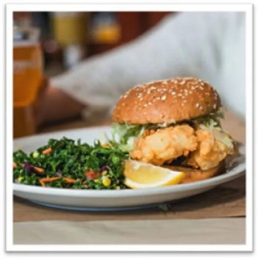 A sesame seed bun sandwich filled with fried chicken and lettuce is served on a white plate with a side of leafy green salad and a lemon wedge. A drink and a persons arm are visible in the blurred background.