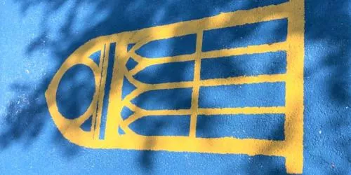 Yellow outline of a stained glass window is painted on a blue ground. Sunlight and tree shadows partially cover the design.