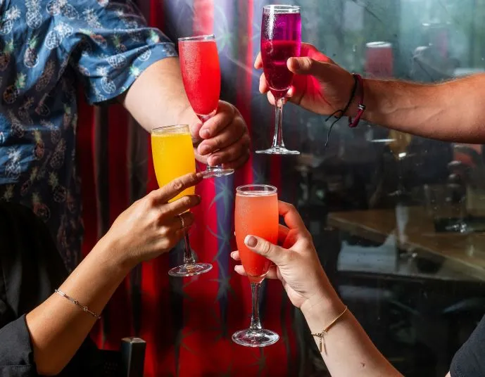 Five hands raise colorful cocktails in tall glasses for a toast indoors, with a patterned red curtain and window in the background. Each drink is a different bright color: red, pink, purple, and orange.