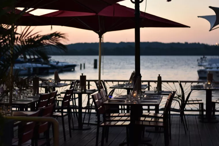 Outdoor restaurant seating with tables and chairs under large red umbrellas, overlooking a river or lake with boats docked nearby at sunset. The scene is calm and softly lit.