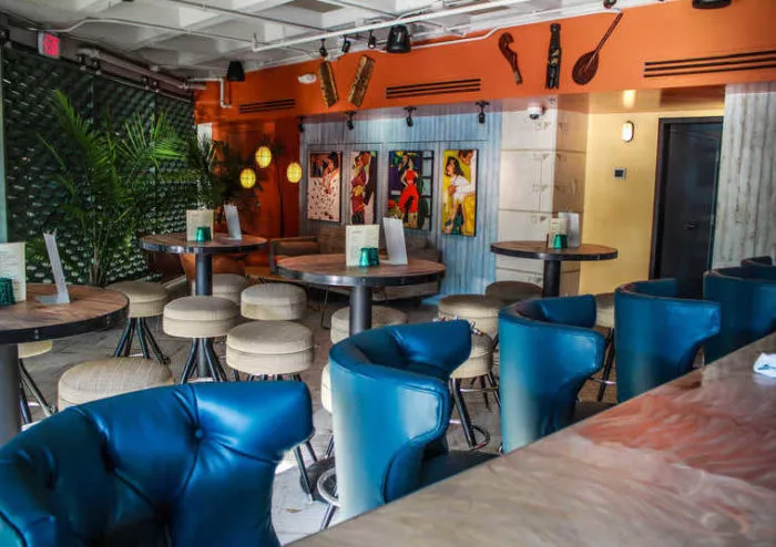 A colorful restaurant interior with teal chairs, round tables, tropical plants, and vibrant artwork on an orange wall, creating a lively and inviting atmosphere. Menus are placed on the tables.