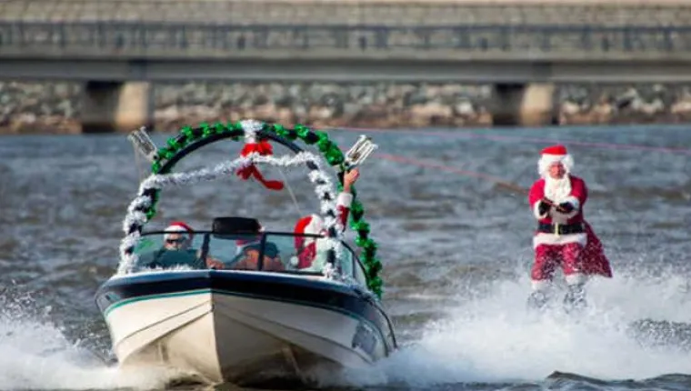 A person dressed as Santa Claus water-skis behind a decorated boat on a river, with another person in a Santa hat driving the boat. A bridge is visible in the background.
